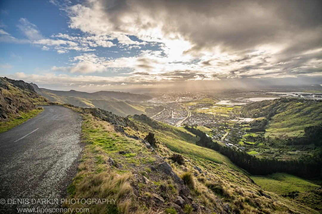 A paved road curves along a grassy ridge toward a distant city and harbor with sunbeams breaking through cloud cover, Central Otago.