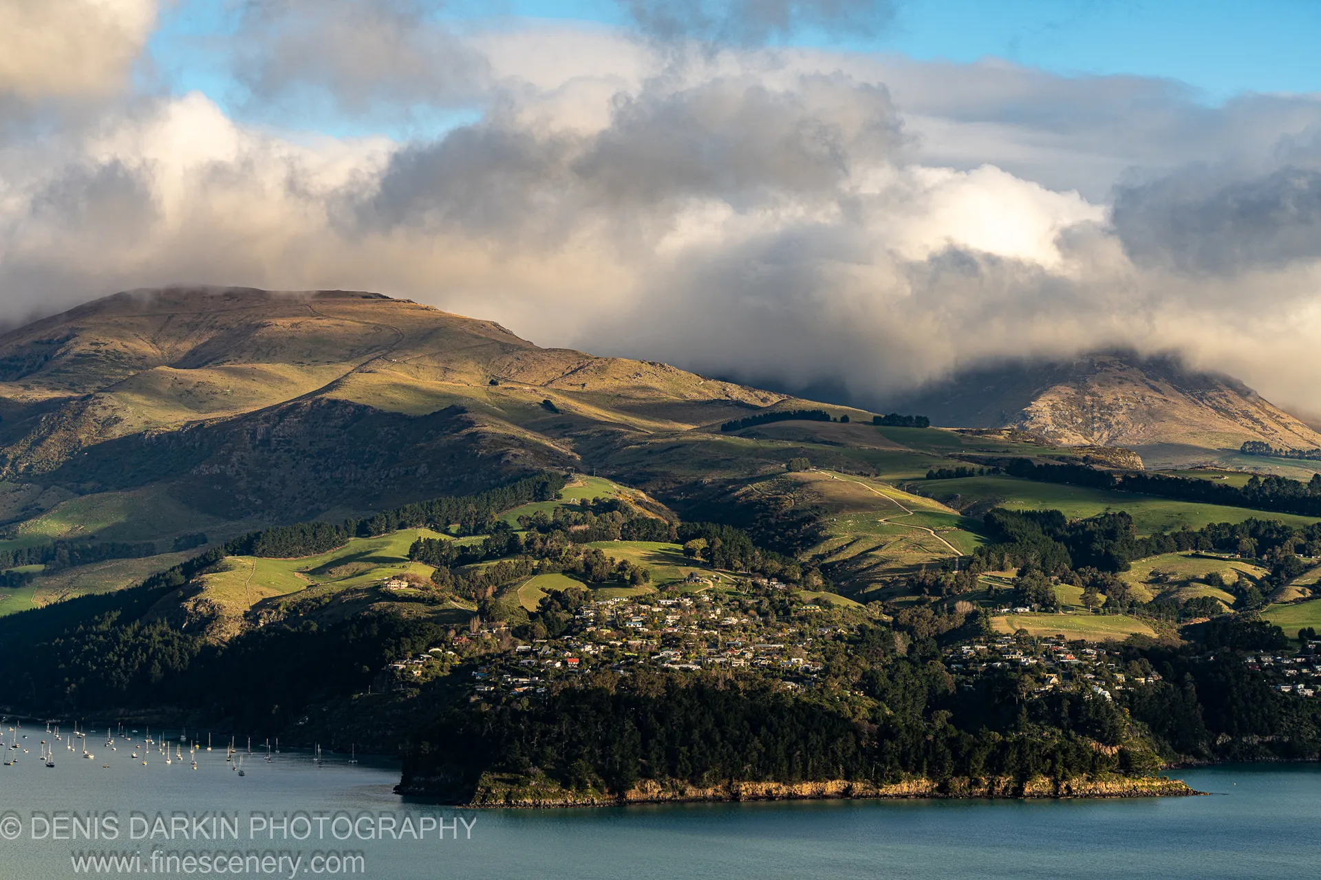 Anchored sailboats on the dark blue lake below mountain ridges in low cloud, with sunlit upper slopes above a lakeside settlement, Central Otago.