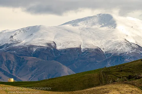 A wooden fence sweeps diagonally across a green hillside toward a snow-covered mountain summit under heavy clouds at dawn, Central Otago, New Zealand.