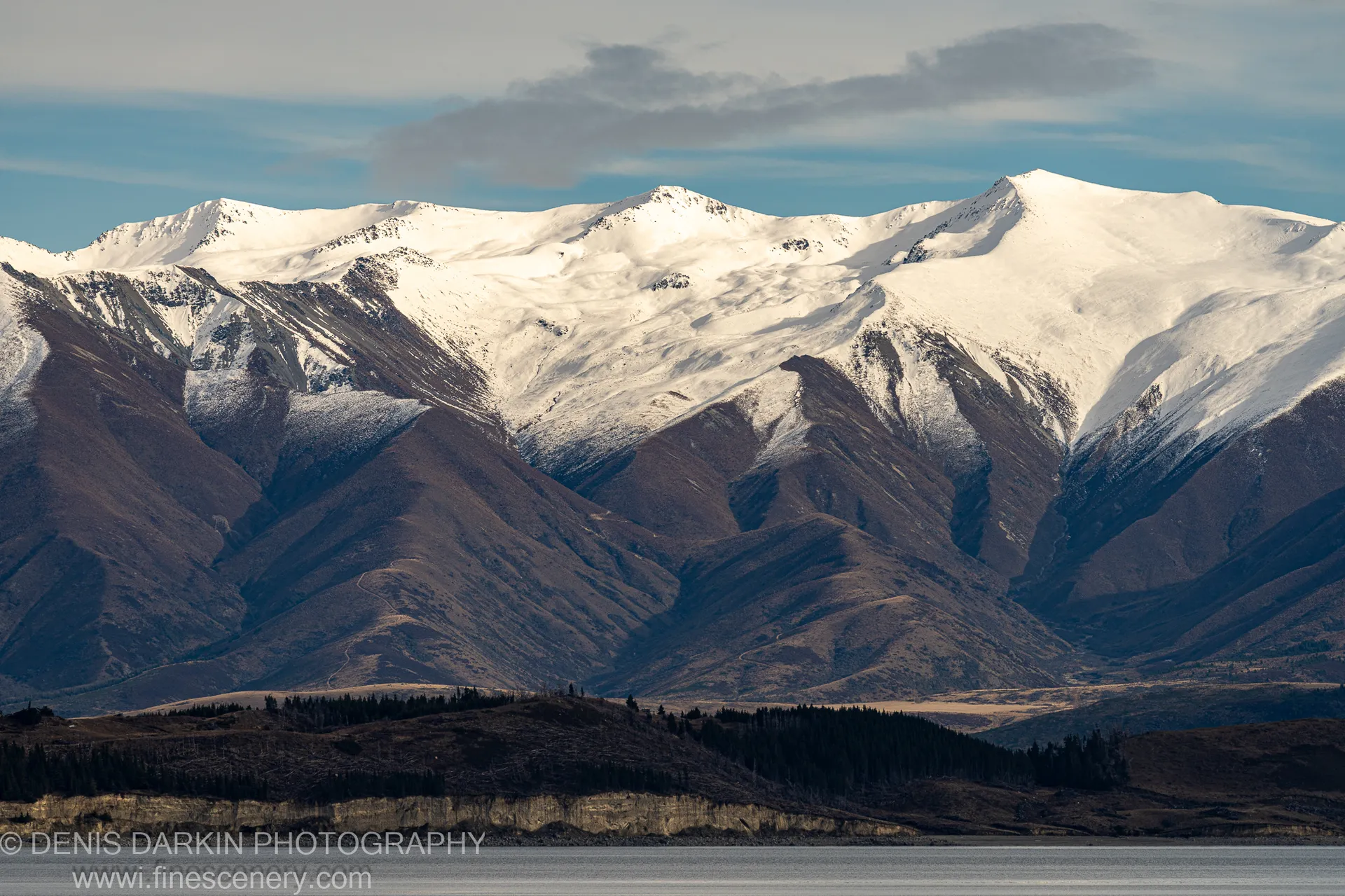 Weathered cliff face with distinct geological strata, a band of coniferous trees at the water's edge, and snow-capped peaks above, Central Otago.