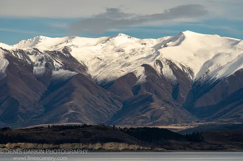 Weathered cliff face with distinct geological strata, a band of coniferous trees at the water's edge, and snow-capped peaks above, Central Otago.