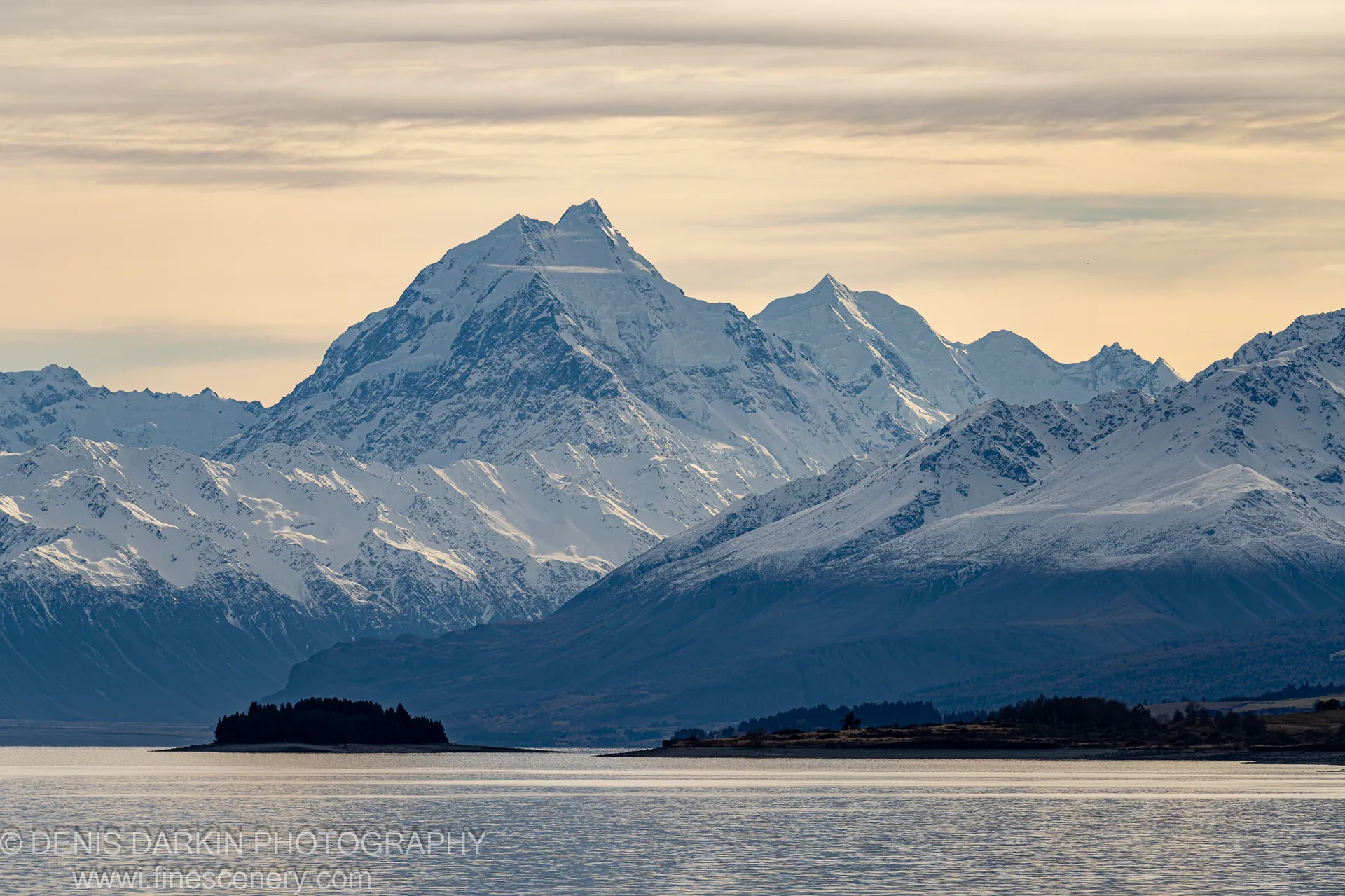 A telephoto view across Lake Wakatipu in winter morning, layered snow-capped peaks and dark forested shorelines receding into pale haze, Queenstown.