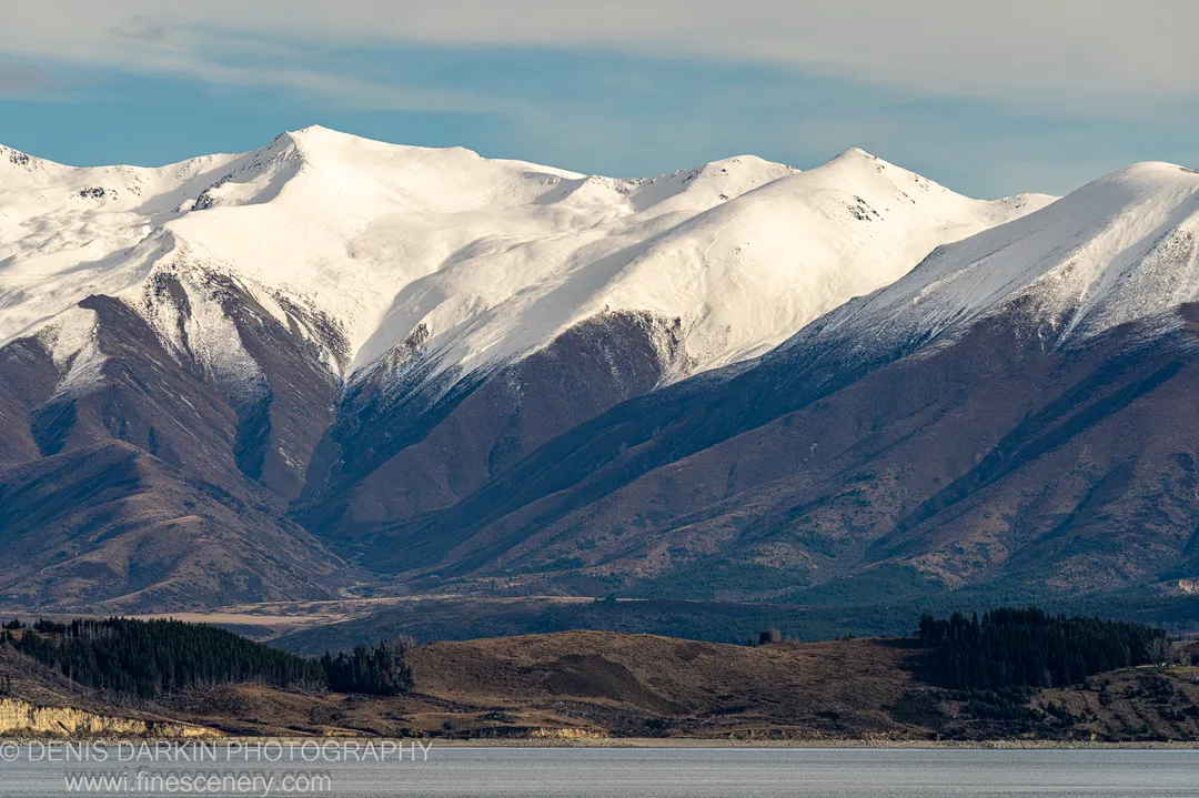 Calm winter lake bordered by dark coniferous forest, with snow-covered mountain ranges rising steeply from the far shore, Central Otago.