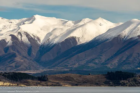 Calm winter lake bordered by dark coniferous forest, with snow-covered mountain ranges rising steeply from the far shore, Central Otago.