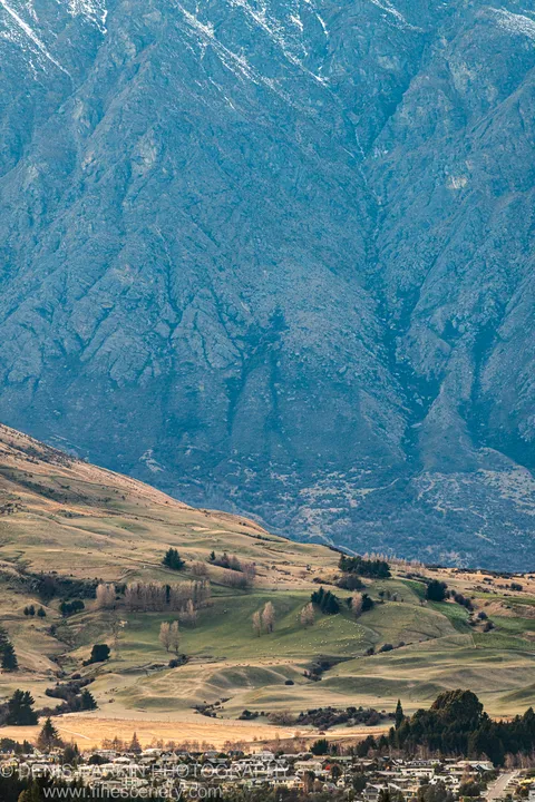 Rolling pasture in predawn light below a towering blue-grey mountain massif, with grazing livestock on the hillside near Queenstown, New Zealand.
