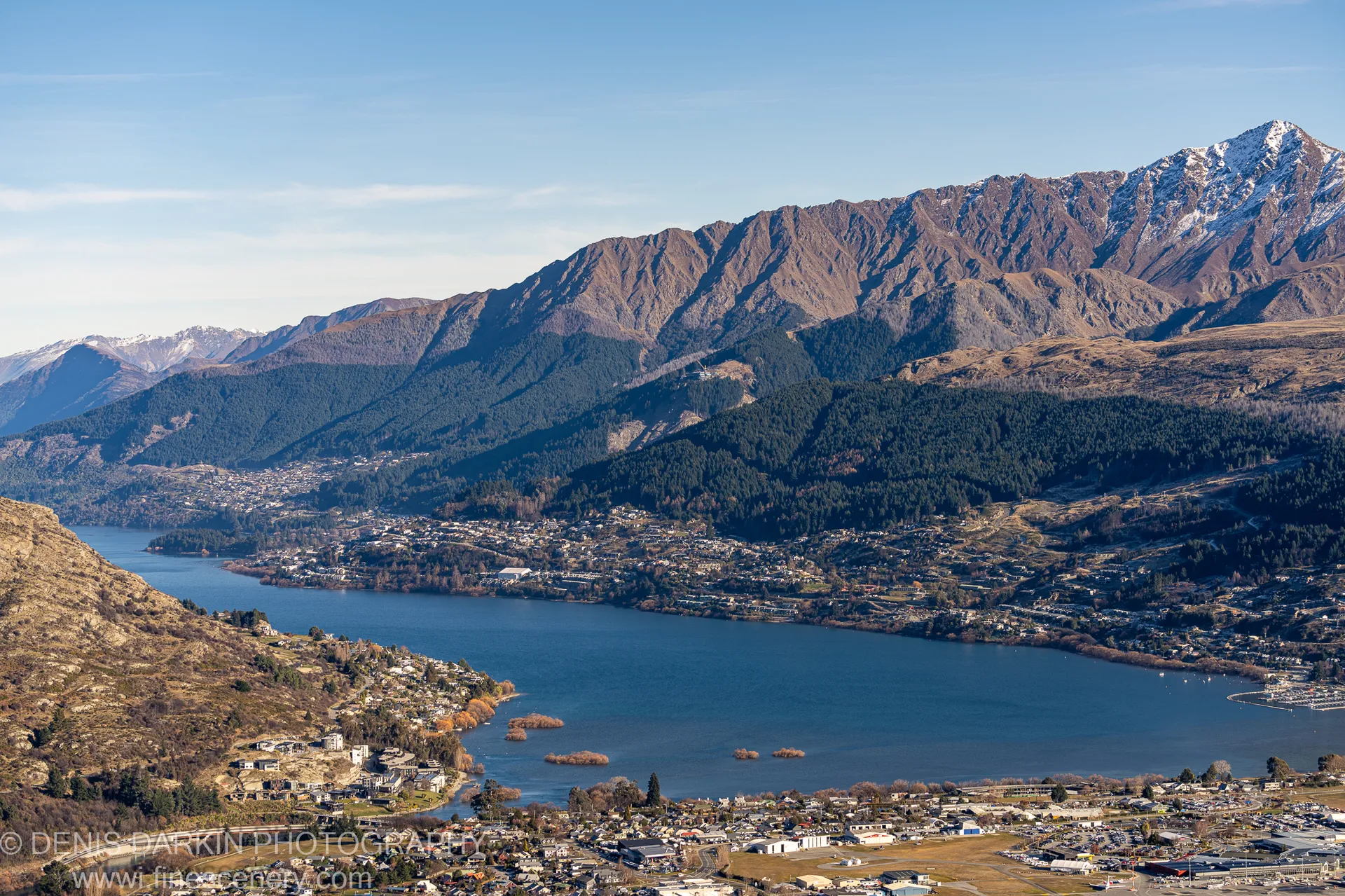 Winter dawn over a mountain-ringed lake at Queenstown, with rocky foreground hills, a town stretching along the shoreline, and snow-capped peaks above.