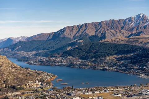 Winter dawn over a mountain-ringed lake at Queenstown, with rocky foreground hills, a town stretching along the shoreline, and snow-capped peaks above.