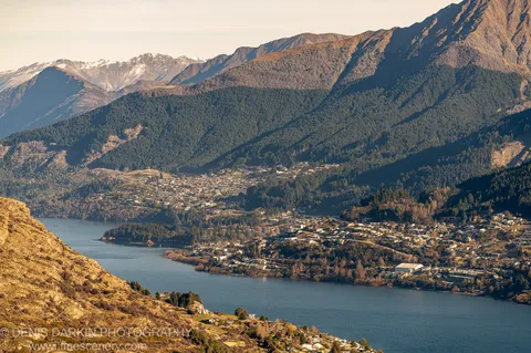 Golden morning light on steep forested hillsides above a blue lake, with long shadows crossing the slopes and a town visible below, Queenstown.