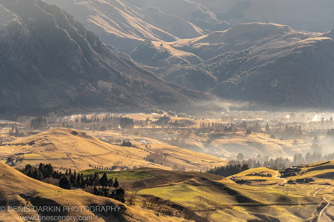 Low morning mist in a valley below sunlit golden-grass hills, with a rural settlement partly obscured by haze, near Queenstown, South Island.