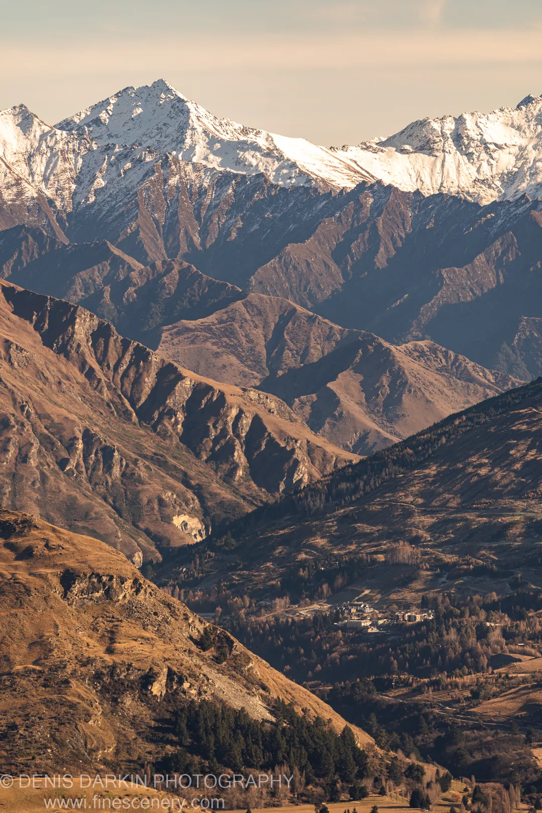 A small valley settlement below dry-grass slopes and layered snow-capped mountain ranges in winter morning light, South Island, New Zealand.