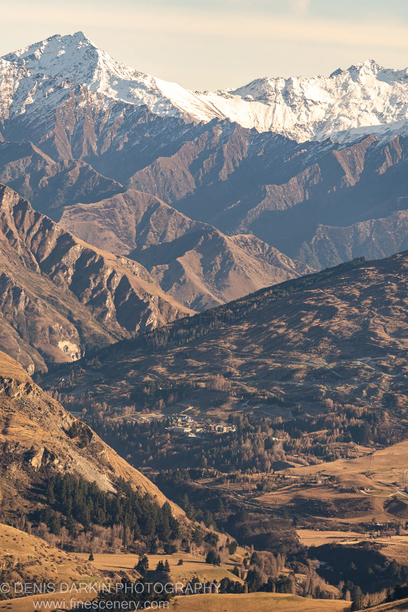 Clustered valley buildings deep in shadow below sunlit snow-dusted mountain ridges, with layered ranges receding into pale morning sky, near Queenstown.