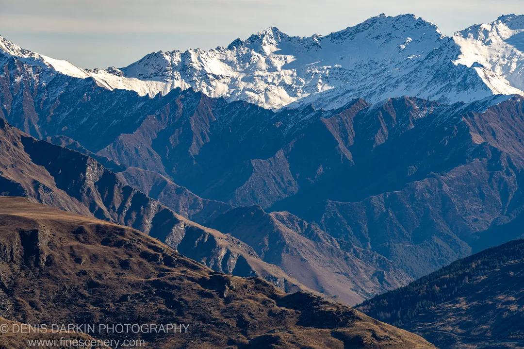 Warm brown foreground mountain slopes above deep blue-shadowed valleys, with jagged snow-capped summits in clear morning light, Queenstown area, South Island.