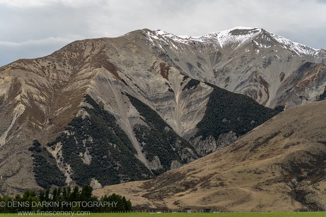 Layered grey and ochre rock ridges descend toward a bright green valley floor below a snow-dusted summit, South Island, New Zealand.