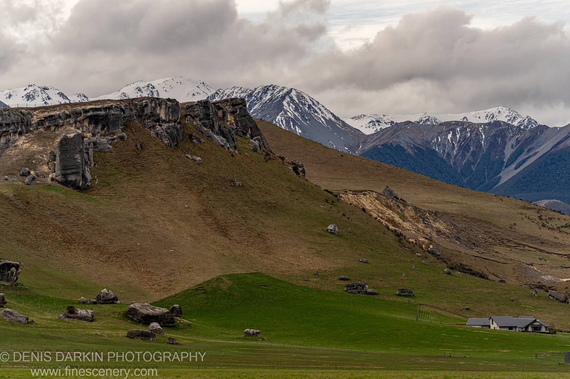 Rolling green slopes scattered with large dark boulders, rising toward snow-capped mountain peaks under a heavy grey cloud bank, South Island, New Zealand.