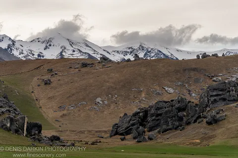 Jagged dark rocks scatter across a slope of green and ochre, with snow-capped peaks rising behind grey cloud, South Island.