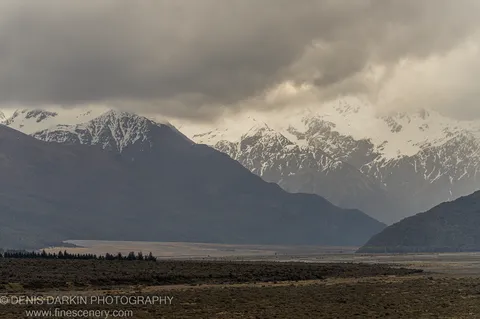 Multiple mountain ranges recede into atmospheric haze beneath a bank of heavy grey cloud, with dark scrub-covered foreground, Punakaiki, South Island, New Zealand.