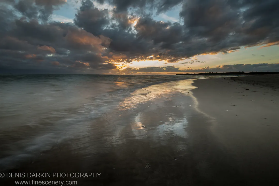 Heavy storm clouds reflected in the wet sand of a flat Fijian beach, with light breaking through at the horizon.