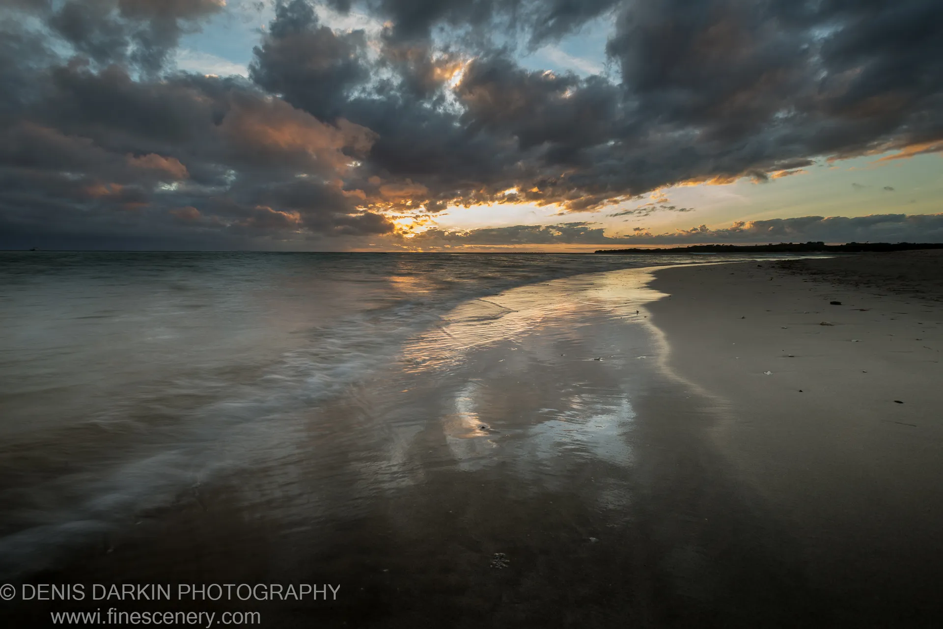 Heavy storm clouds reflected in the wet sand of a flat Fijian beach, with light breaking through at the horizon.