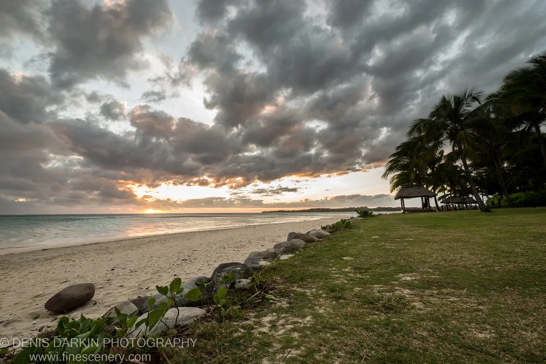 A thatched gazebo sheltered by leaning coconut palms on a rocky Fijian beach, with storm clouds massing over the ocean beyond.