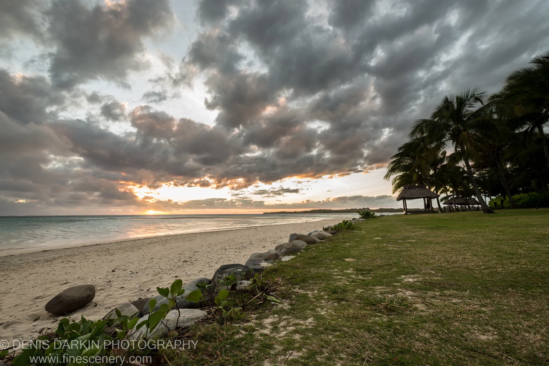 A thatched gazebo sheltered by leaning coconut palms on a rocky Fijian beach, with storm clouds massing over the ocean beyond.