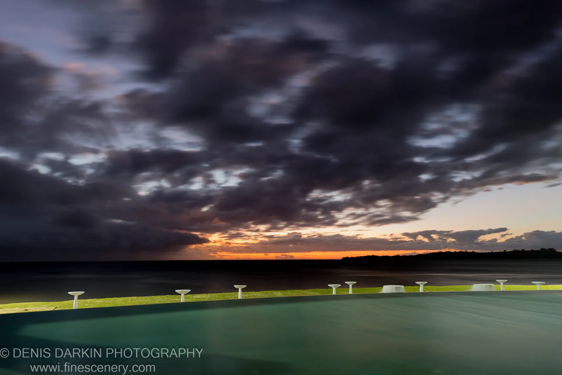 A calm aquamarine pool near the ocean reflects cloud motion and a golden horizon, bordered by white concrete bollards receding into the distance.