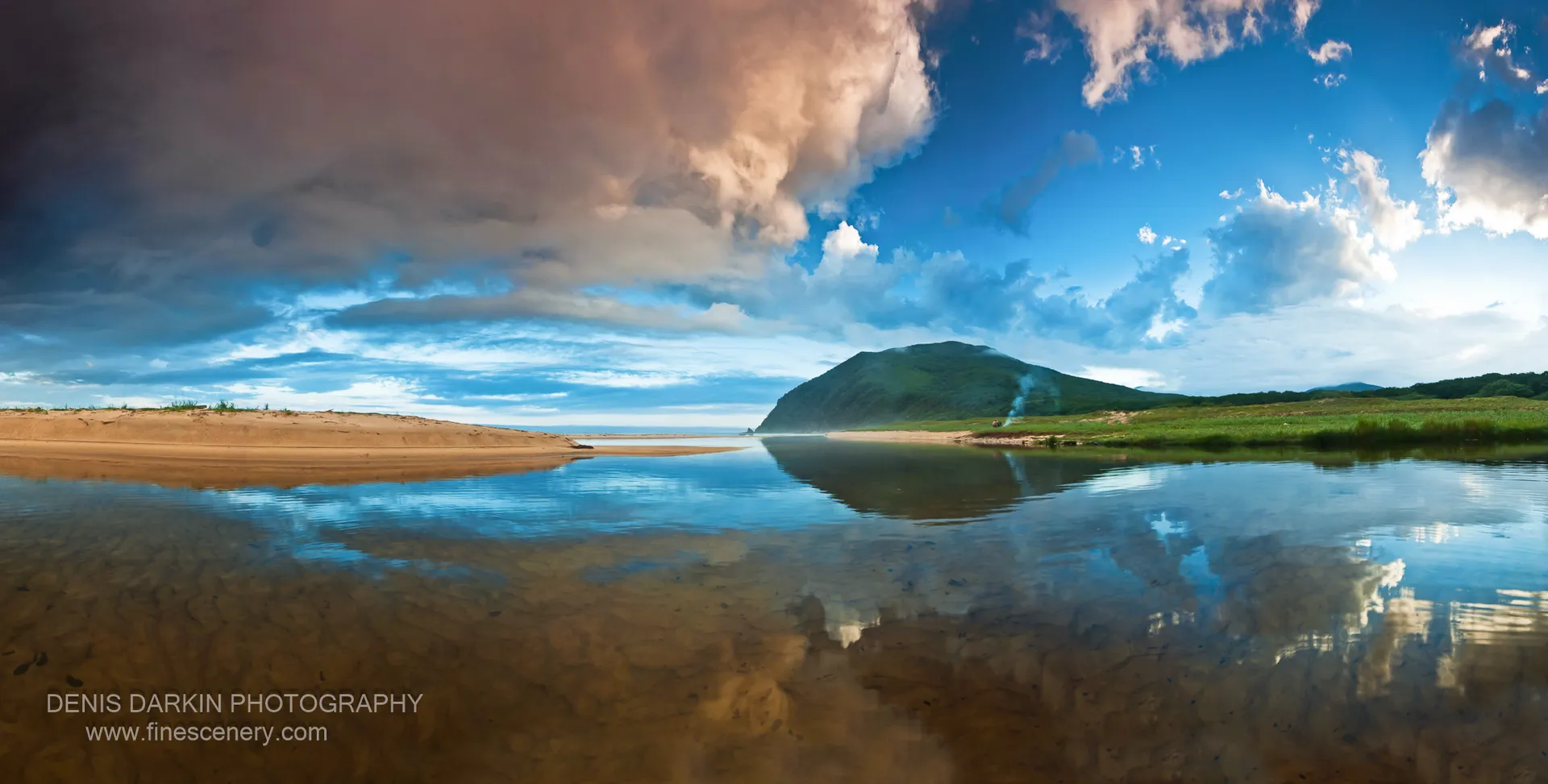 A rounded green hill reflected in mirror-flat water beside a sandy spit at Tachingouza Bay, with tall cumulus clouds in the evening sky, July 2010.