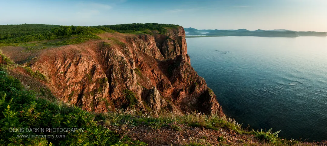 Warm morning light strikes the reddish-brown layered cliff face at Vladimir Bay, above the calm blue waters of the bay below.