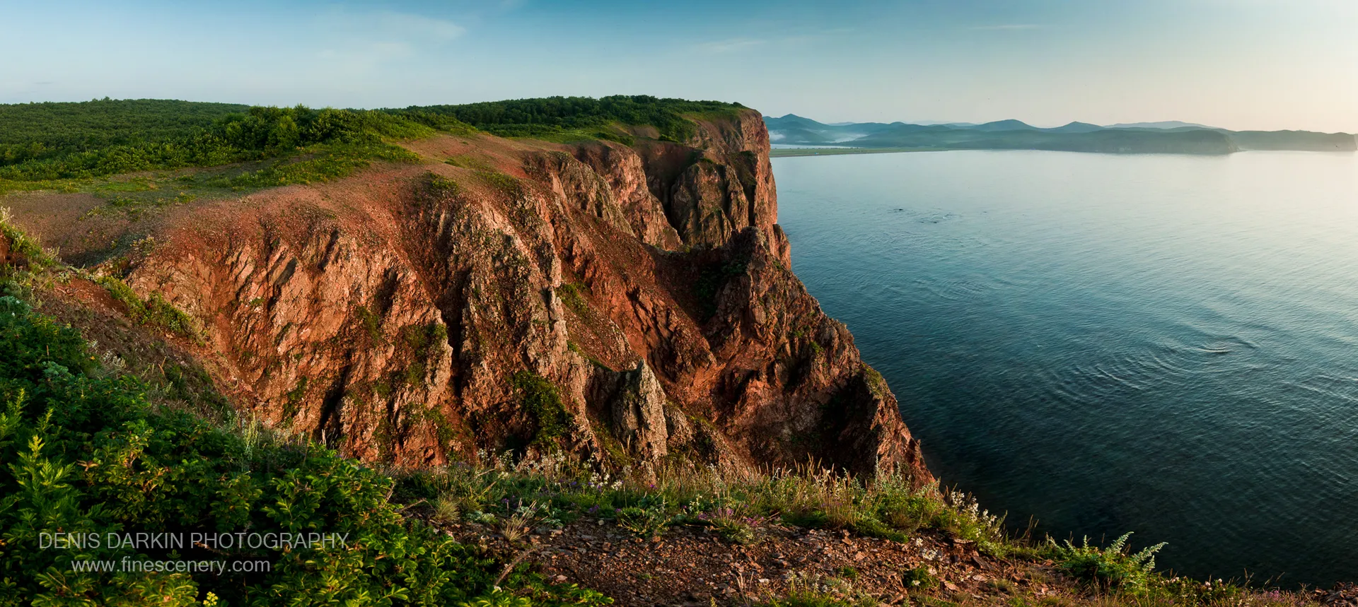 Warm morning light strikes the reddish-brown layered cliff face at Vladimir Bay, above the calm blue waters of the bay below.