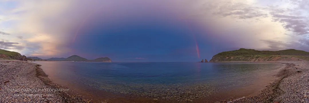A curving pebble beach leads toward distant green headlands and sea stacks under purple-toned clouds in Primorsky Krai.