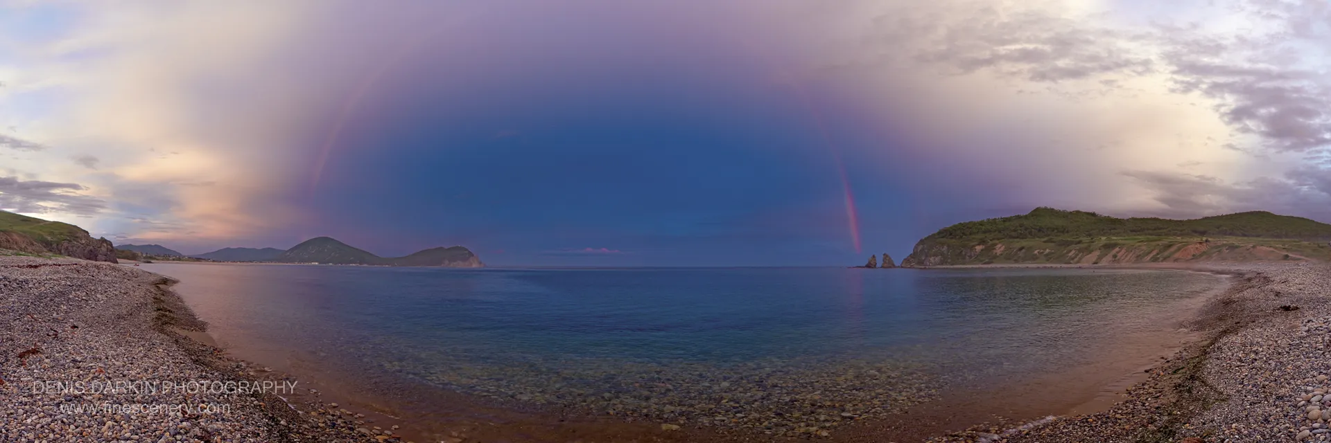 A curving pebble beach leads toward distant green headlands and sea stacks under purple-toned clouds in Primorsky Krai.