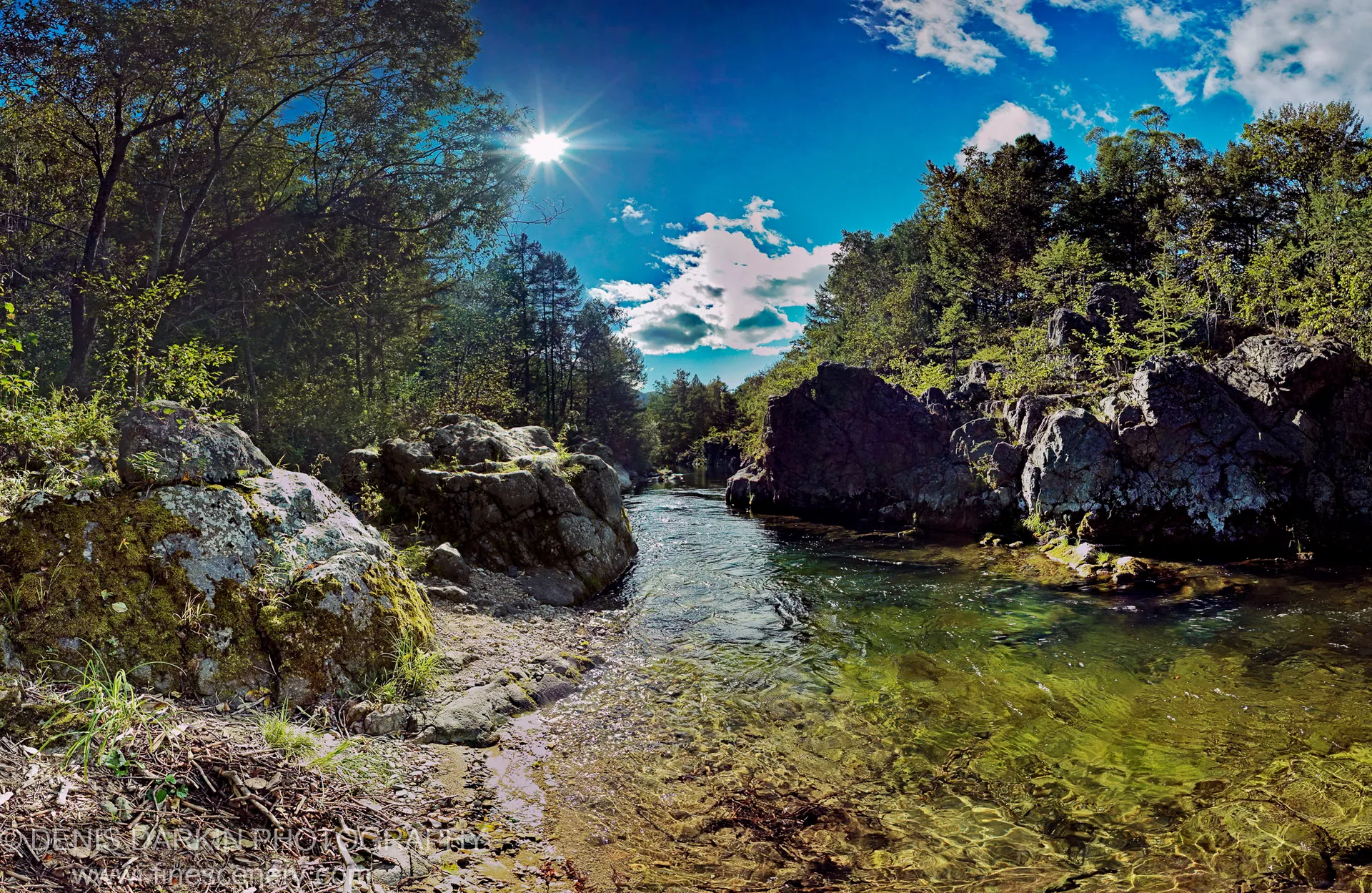 Clear water flows through a rocky taiga gorge in the Russian Far East, with lichen-covered boulders and dense conifer forest as sunlight bursts from above.