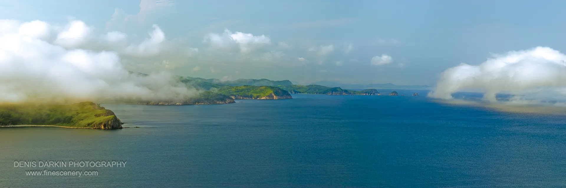 Rocky cliffs and a green-topped peninsula overlook a calm bay at Hasan, Russian Far East, with sea fog softening the distant headlands.