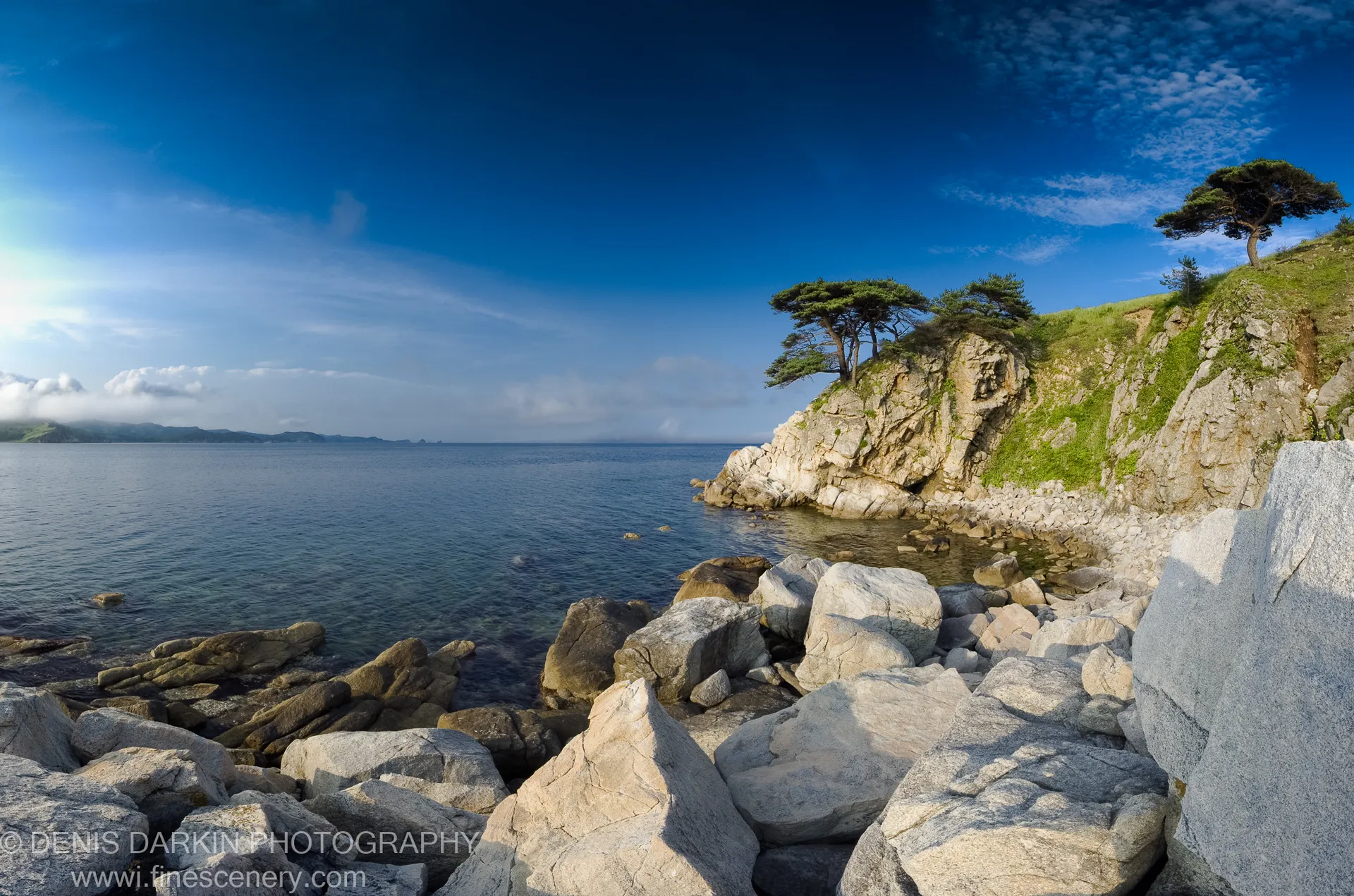 Sun-lit granite boulders on a rocky shoreline at Astafyeva Bay, with a pine-topped headland and calm blue waters beyond.
