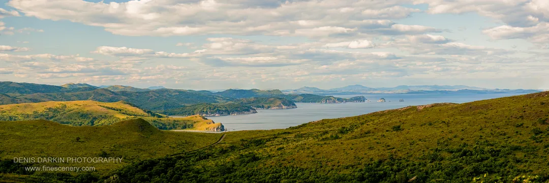 Grassy rolling hills curve toward a calm bay at Gamov Cape under a sky of scattered cumulus, with rocky headlands and islands beyond.