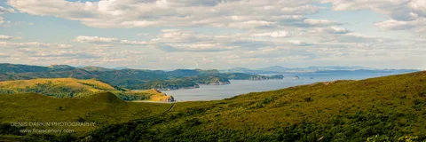 Grassy rolling hills curve toward a calm bay at Gamov Cape under a sky of scattered cumulus, with rocky headlands and islands beyond.