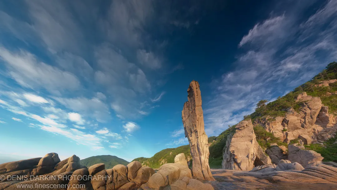 A slender granite spire stands at the center of the Tachingouza Bay coastline, with smooth boulders in the foreground and pines on the hillside.