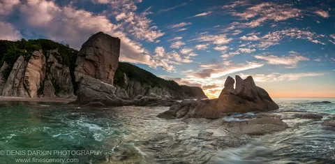 Long-exposure waves wash over smooth coastal stones at Tachingouza Bay, with granite sea stacks and low cliffs visible through the morning mist.