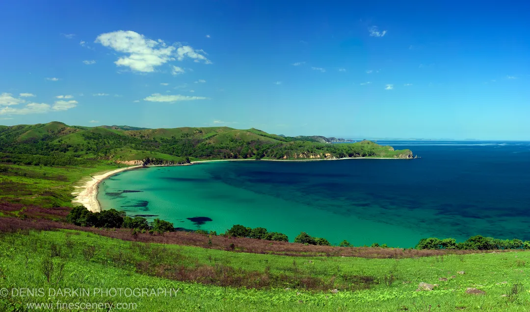 A horseshoe bay of deep blue water and sandy beach at Astafyeva Bay, Far East Marine Reserve, with forested headlands and scattered clouds above distant islands.