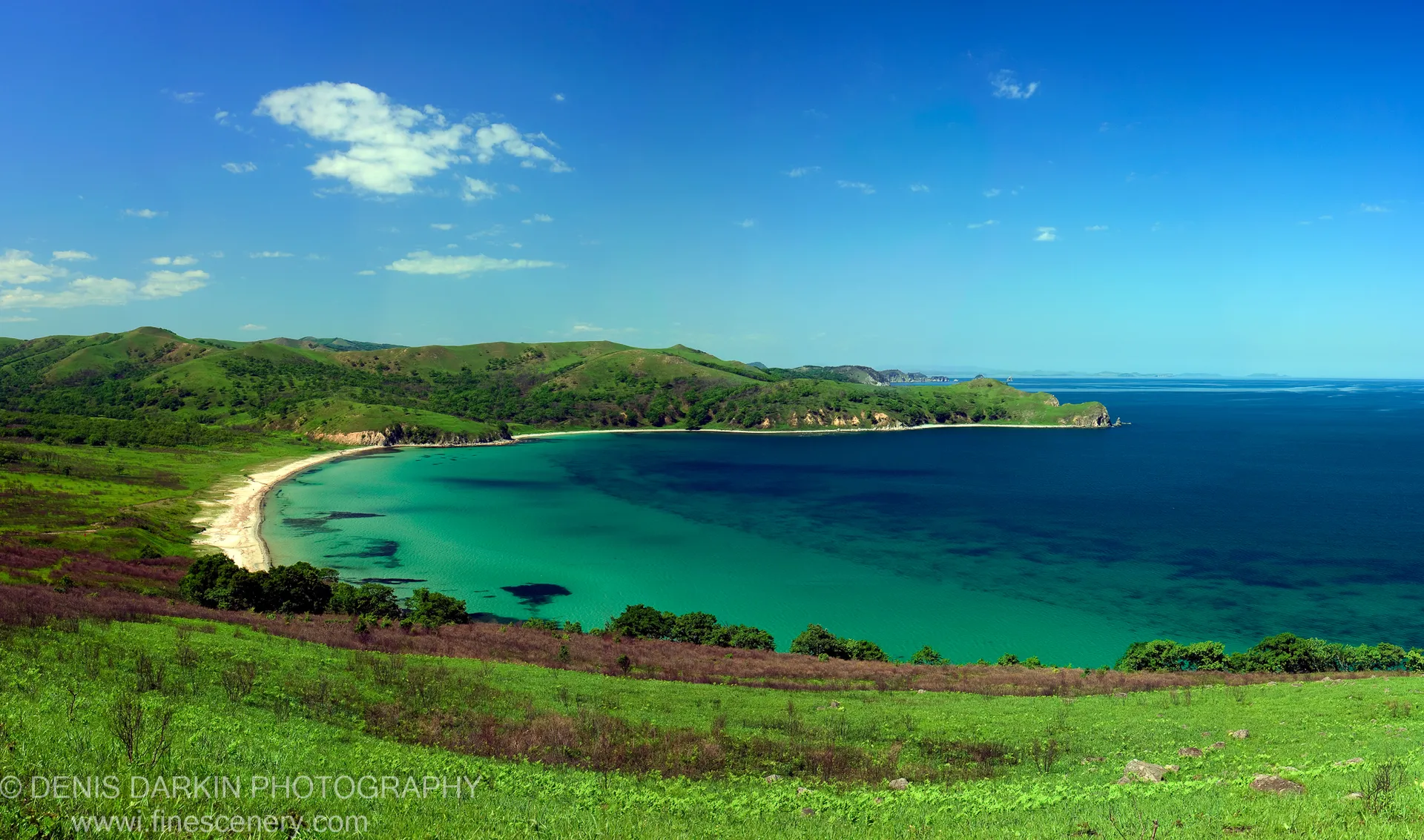 A horseshoe bay of deep blue water and sandy beach at Astafyeva Bay, Far East Marine Reserve, with forested headlands and scattered clouds above distant islands.