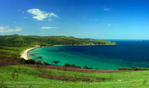 A horseshoe bay of deep blue water and sandy beach at Astafyeva Bay, Far East Marine Reserve, with forested headlands and scattered clouds above distant islands.