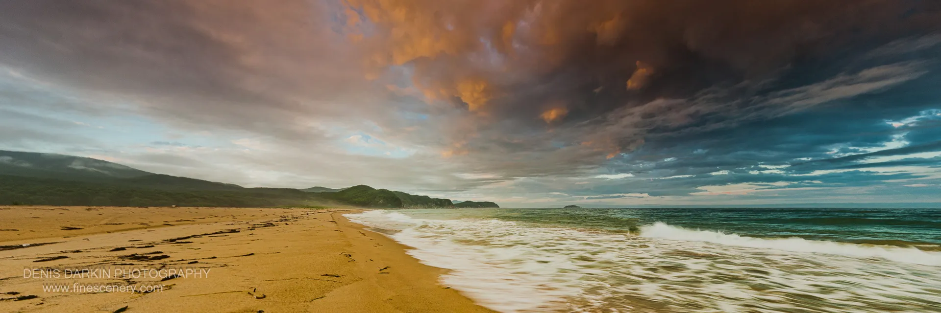 Dark driftwood logs on warm golden sand at Tachingouza Bay, with textured storm clouds catching the last of the evening light overhead, July 2010.