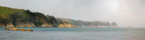 Warm sunlit boulders anchor the left foreground as forested cliffs and pale rolling hills recede into a hazy horizon above calm seas.