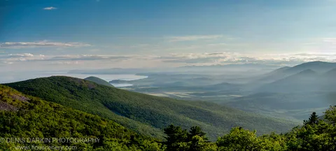 Multiple layers of haze-blue mountain ridges recede toward a distant bay below Kamenistyaya Mountain, Primorsky Krai, under a pale cloud-streaked sky.