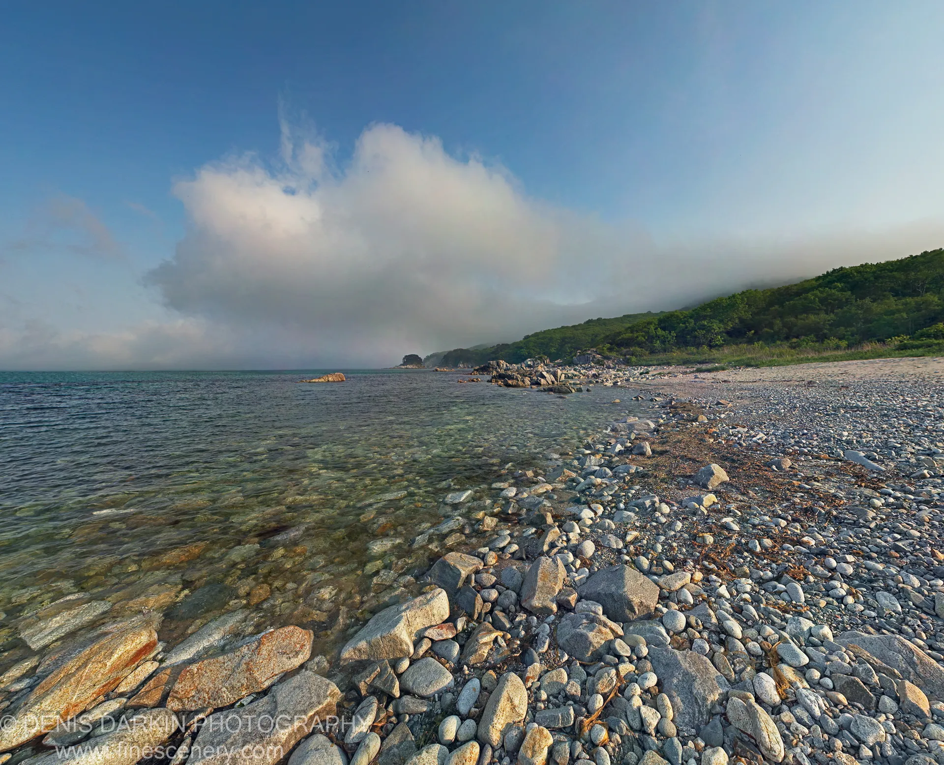 A pebble shoreline at Telyakovskogo Bay curves toward a forested headland under a large cumulus cloud in a blue sky.