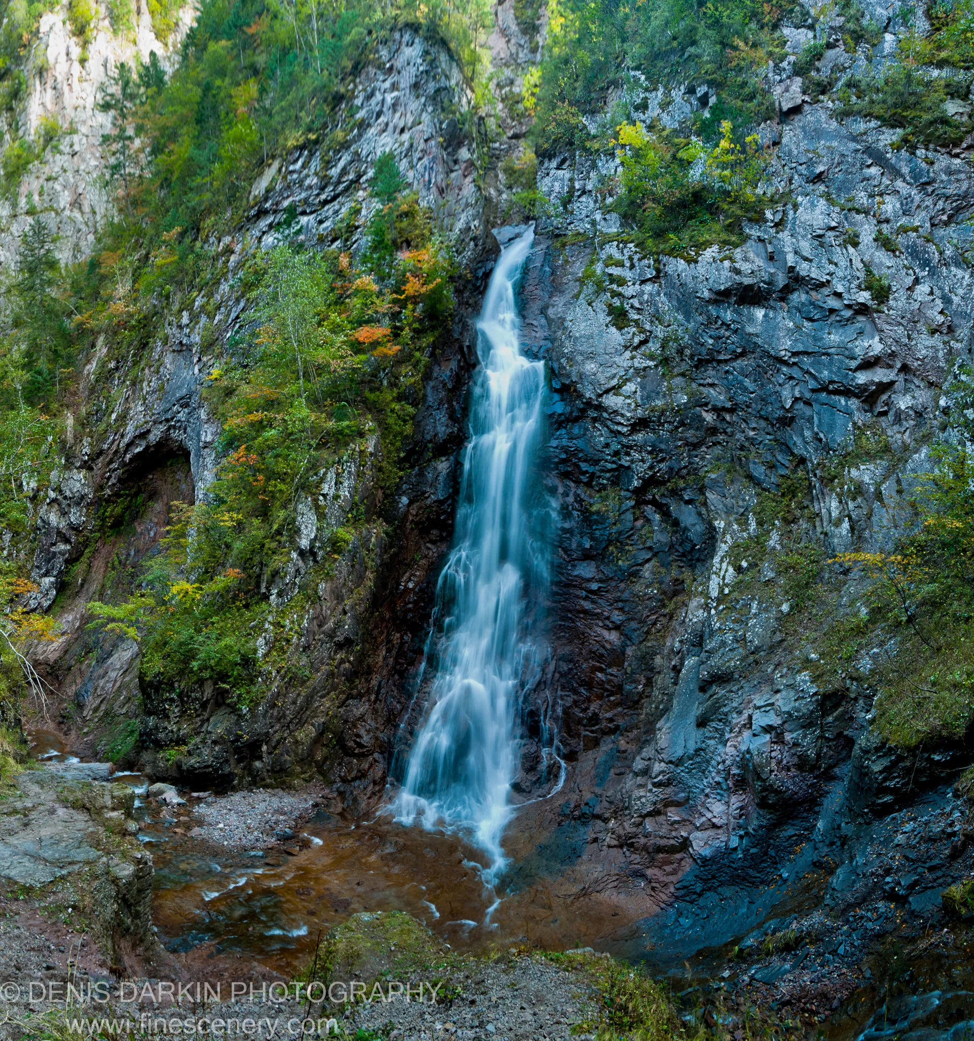 A slender waterfall drops down a grey rock face into a pool, with green moss and orange autumn foliage, Black Shaman Waterfall.