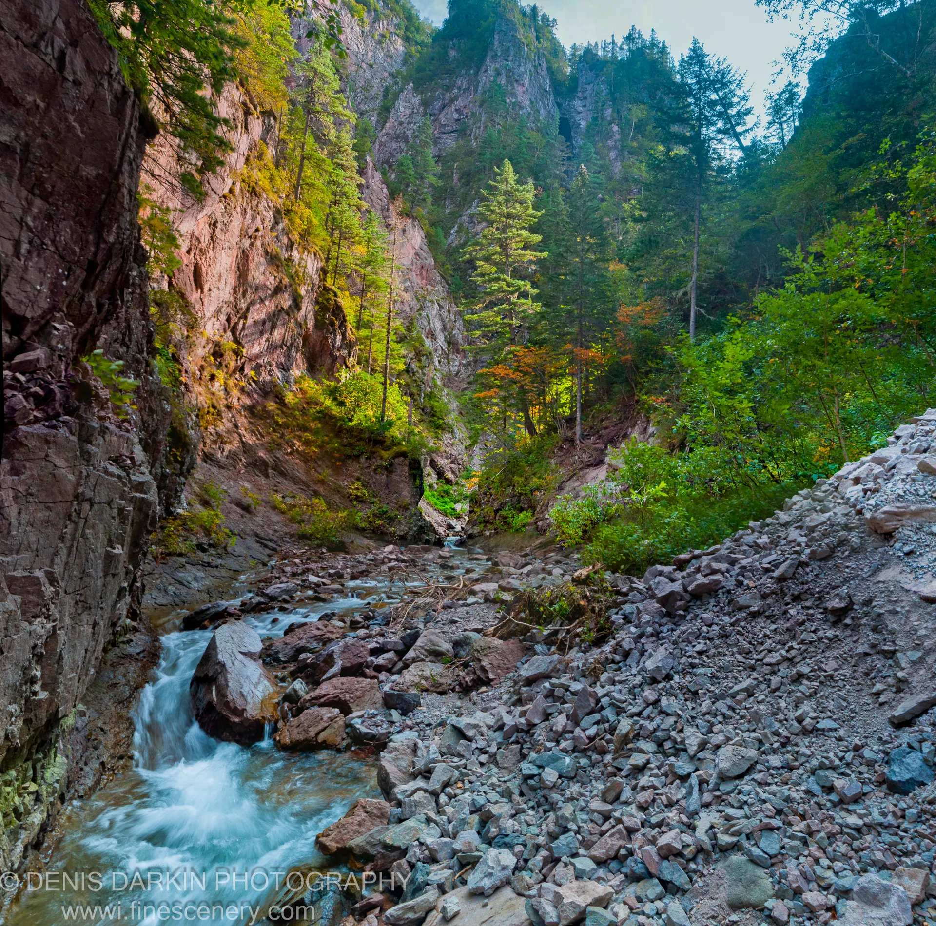 A rushing stream flows through a narrow rocky gorge with a gravelly bank and steep cliffs above, orange autumn foliage visible, Terneysky District.