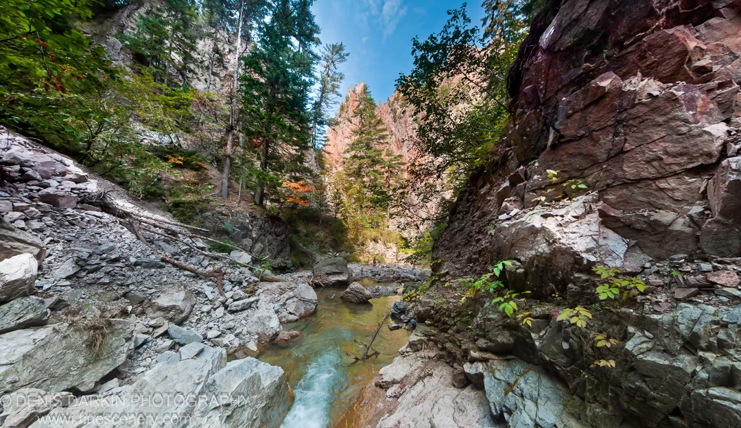 Sunlit stratified cliffs above a shallow river flowing through smooth stones, with coniferous trees and autumn color, Terneysky District.