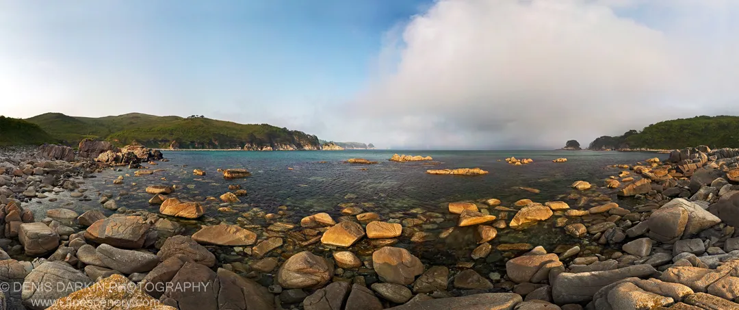 Rounded boulders catch morning light where shallow water meets the cove shore, with forested headlands and a cloud formation in the background.