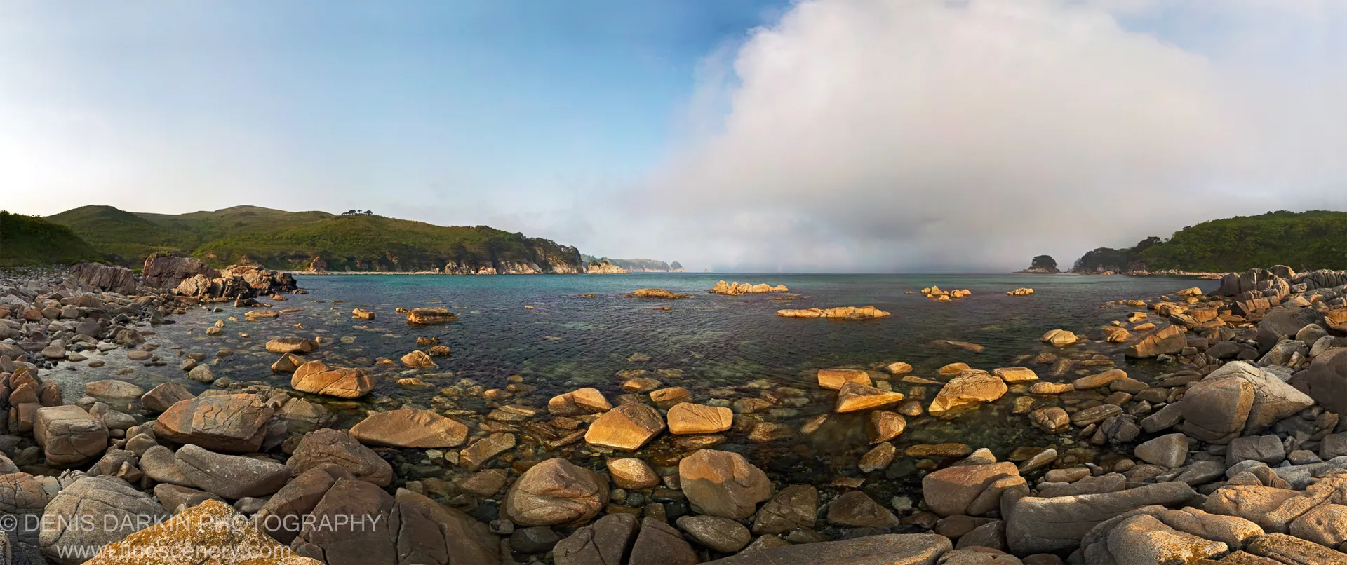 Rounded boulders catch morning light where shallow water meets the cove shore, with forested headlands and a cloud formation in the background.
