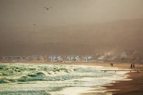 Turbulent green waves breaking in the foreground before a row of blue-roofed beach cabins at the base of a fog-shrouded hill, Shamora Bay.